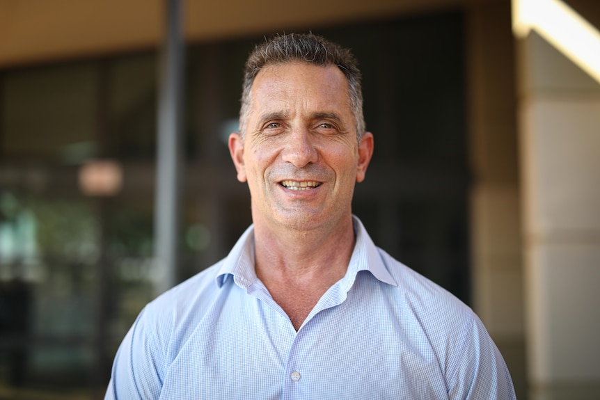 A man in a light blue collared shirt smiles at the camera 