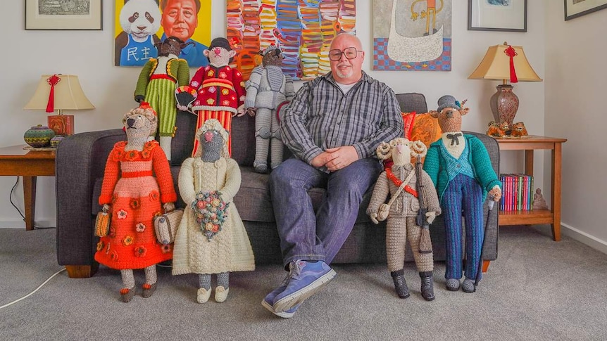 A man in jeans and a button-up shirt with large glasses sits on a couch surrounded by metre-tall crochet characters.