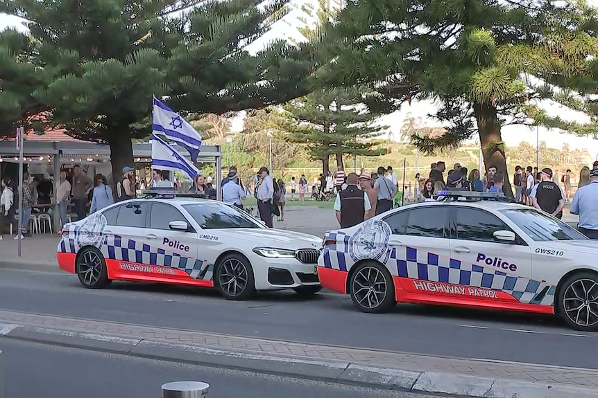two police cars patrol coogee beach as people stand along the curb carrying israeli flags ahead of a pro-palestinian convoy