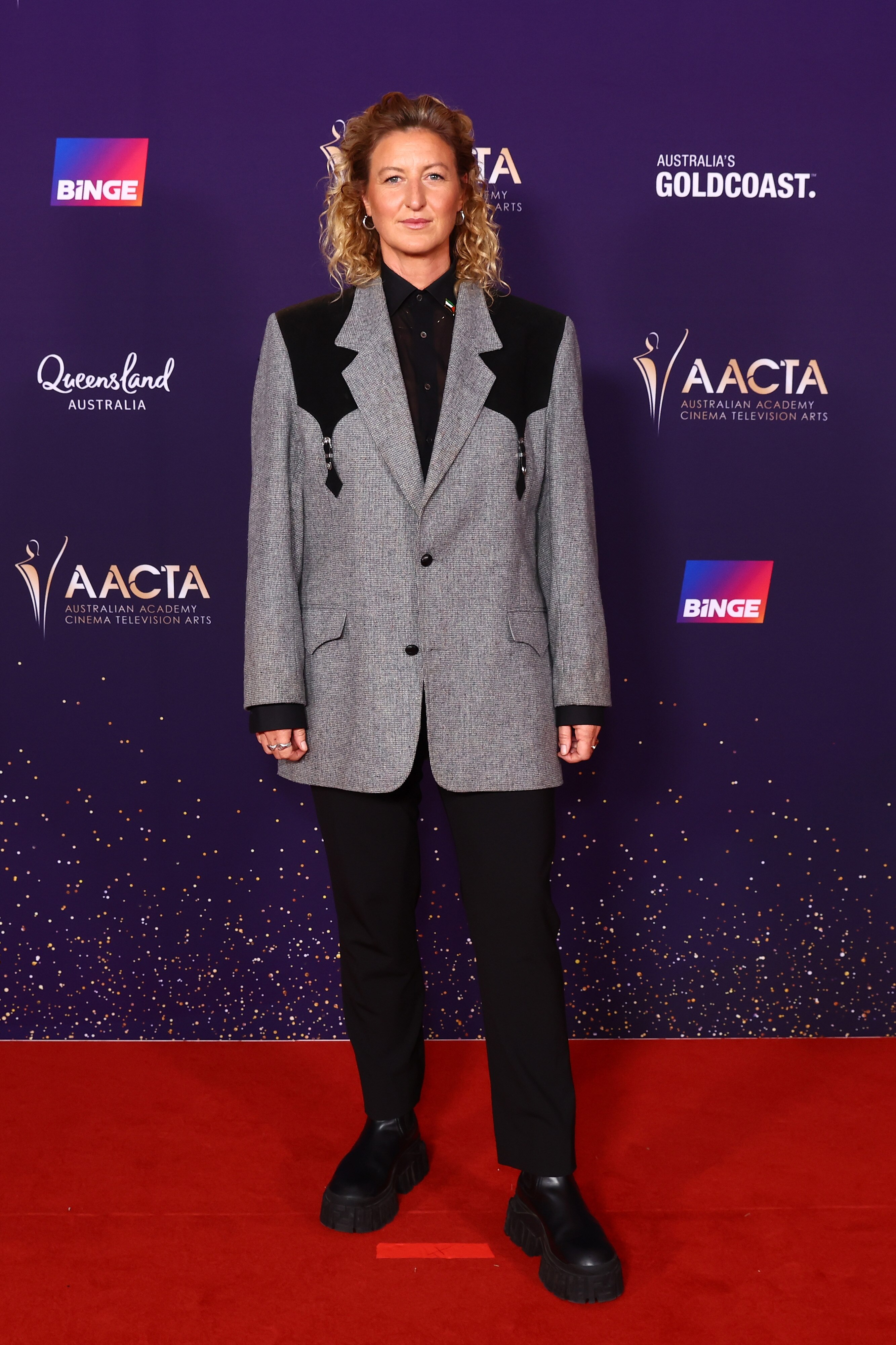 Kate Box wears a black and grey suit jacket with a black shirt, black trousers and black chunky shoes, hair down but swept back