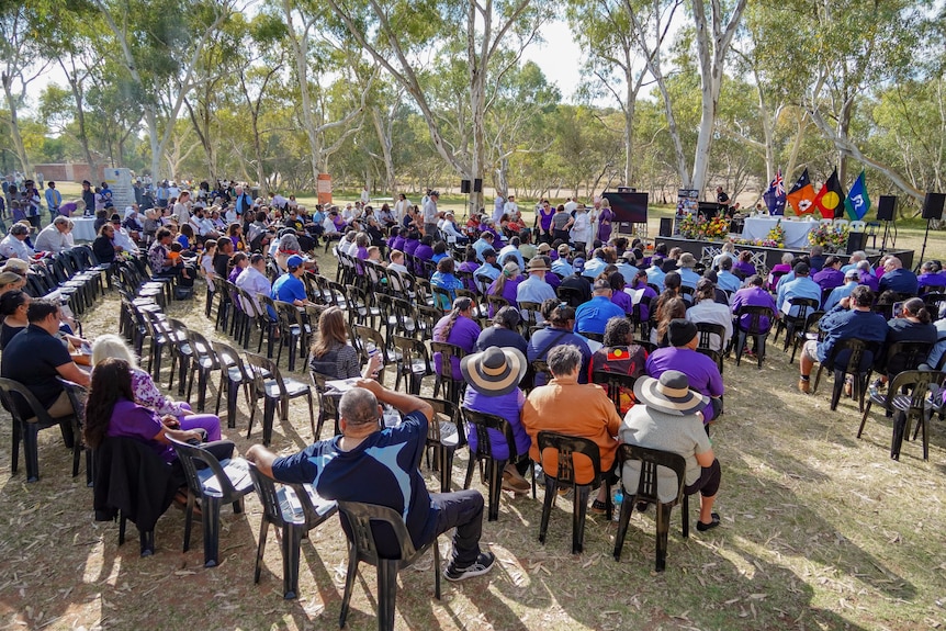 A crowd of people sit in plastic chairs facing a small stage. Many are wearing purple and blue