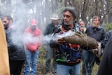 An Aboriginal man in a patterned jumper and jeans holds a smoking burial parcel, surrounded by his Anaiwan peers