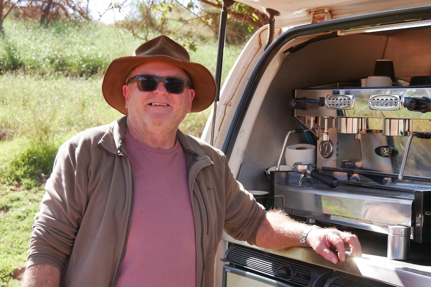 A man wearing sunglasses and a hat stands next to a coffee machine built into the back of a van.