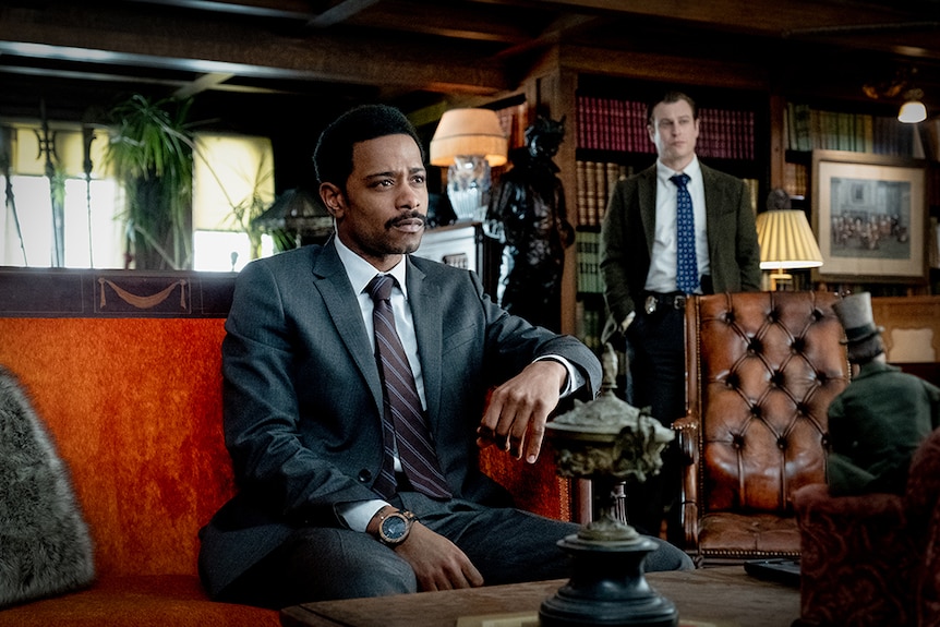 LaKeith Stanfield in suit sits on orange red lounge, Noah Segan stands behind, both in ornate interior sitting space with books.