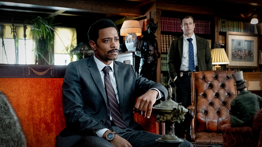 LaKeith Stanfield in suit sits on orange red lounge, Noah Segan stands behind, both in ornate interior sitting space with books.