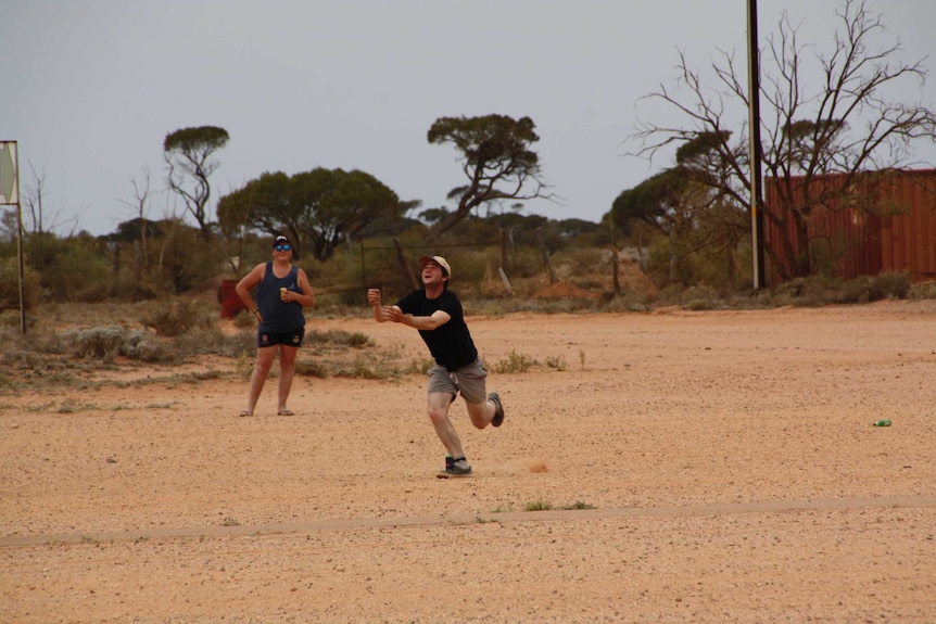 A man in a black shirt and yellow cap runs to catch a falling ball while another man drinks a beer.