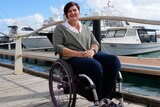 A woman in a wheelchair smile at the camera while sitting on a boardwalk with a boat in the background