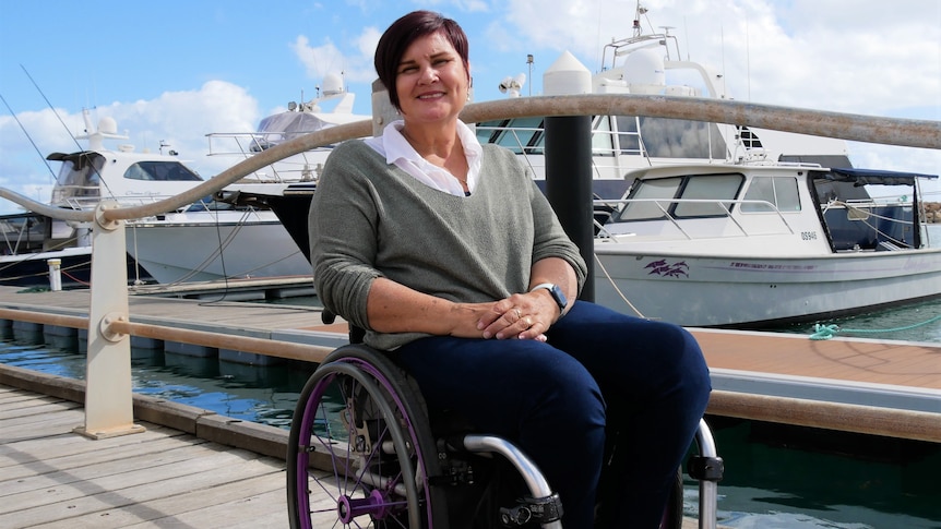 A woman in a wheelchair smile at the camera while sitting on a boardwalk with a boat in the background