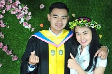 A man wearing a graduation gown with his arms around a woman with black hair giving a thumbs up.