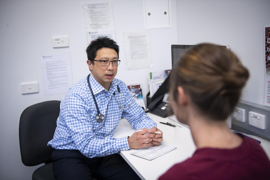 Jason Tye-Din, sitting at desk in check shirt, with stethoscope around neck, talking to person blurred and visible from behind.