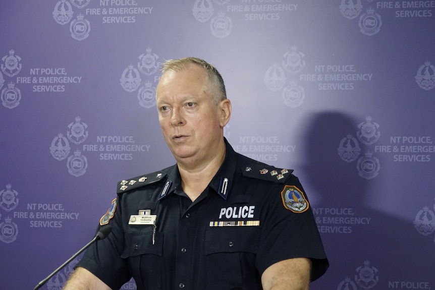 A man in a NT Police uniform standing at a lectern and speaking, in front of a  purplebanner featuring the NT Police logo .