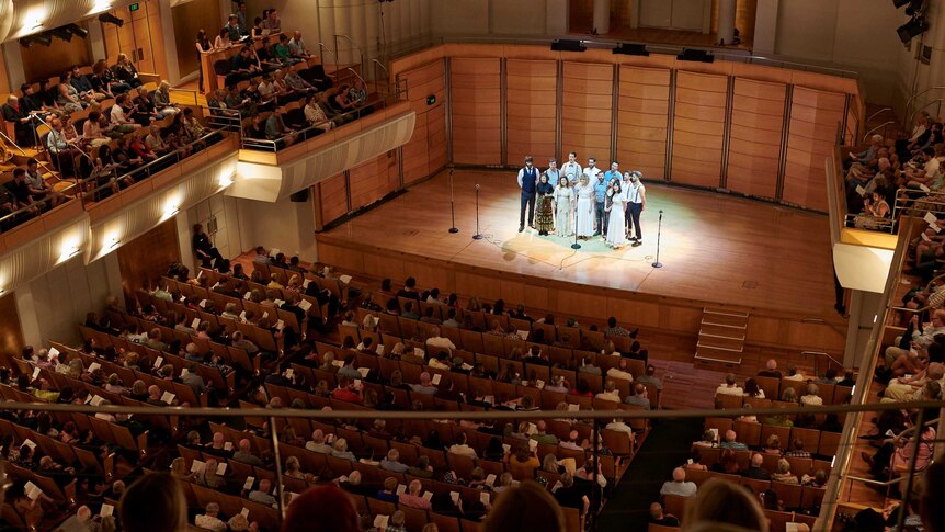 Musicians stand together on stage in front of a packed auditorium.