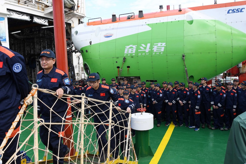 A crew of uniformed submariners calmy disembark a ship onto a dock, passing a huge green submarine.