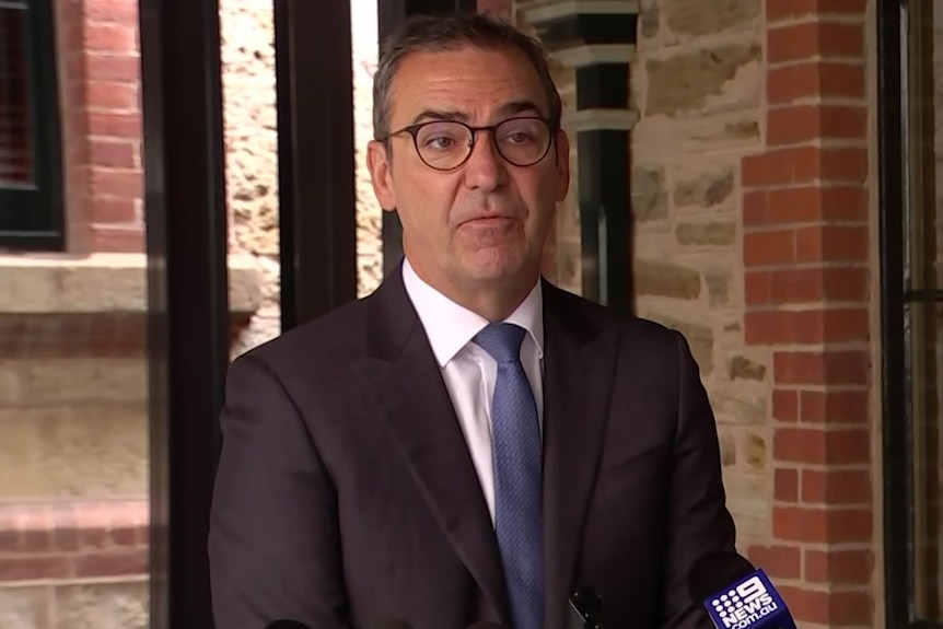 A man wearing a black suit, white shirt, purple tie and black glasses stands behind microphones in front of a brick building