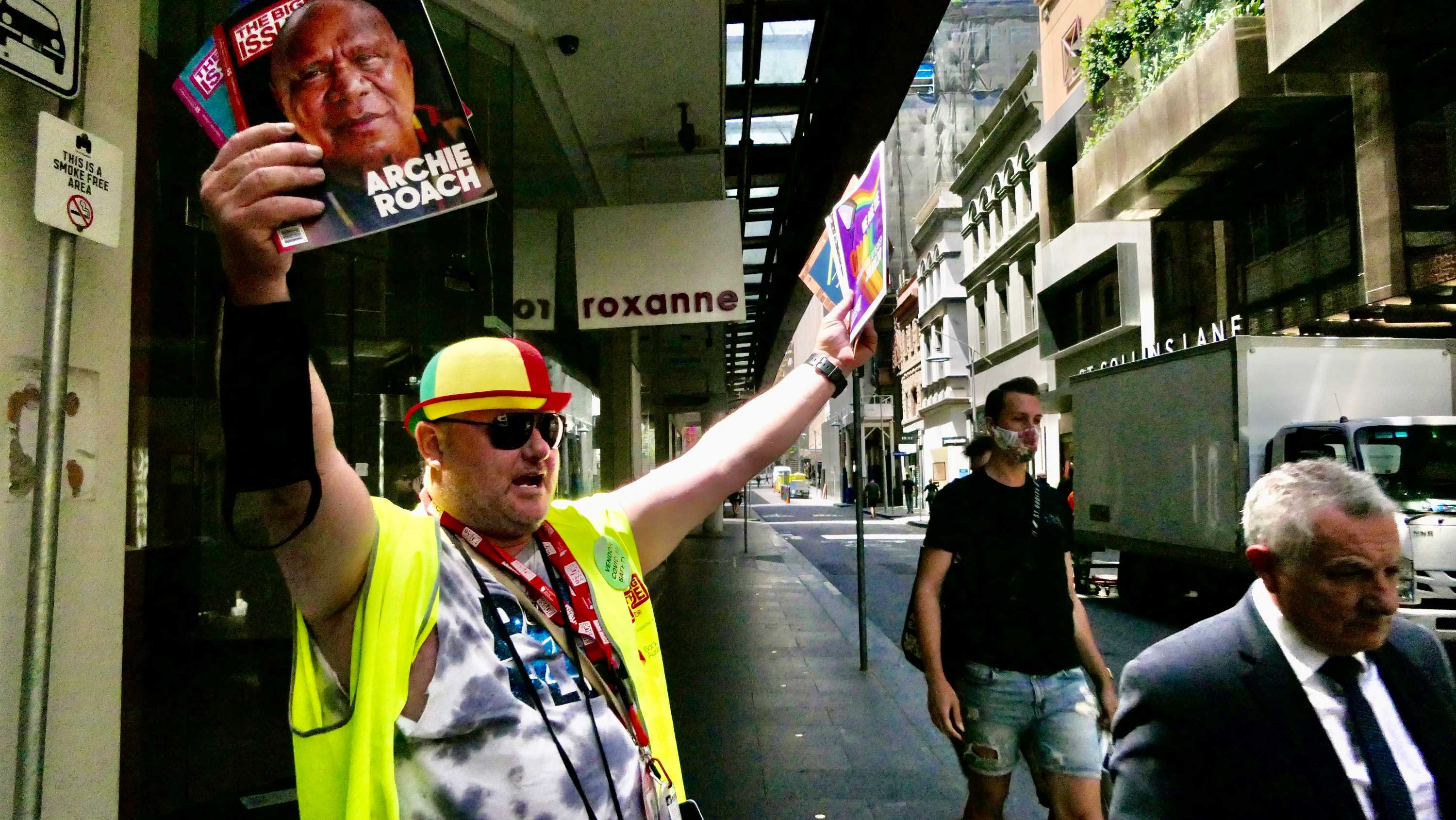 A Big Issue vendor dressed in a fluro vest and hat spruiking the magazine on the street.