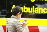 A woman wearing a blue surgical mask and using a phone next to an ambulance.