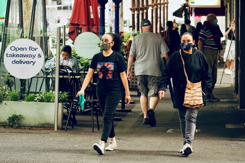 Two women wearing black jeans and face masks walk past a cafe on a city street.