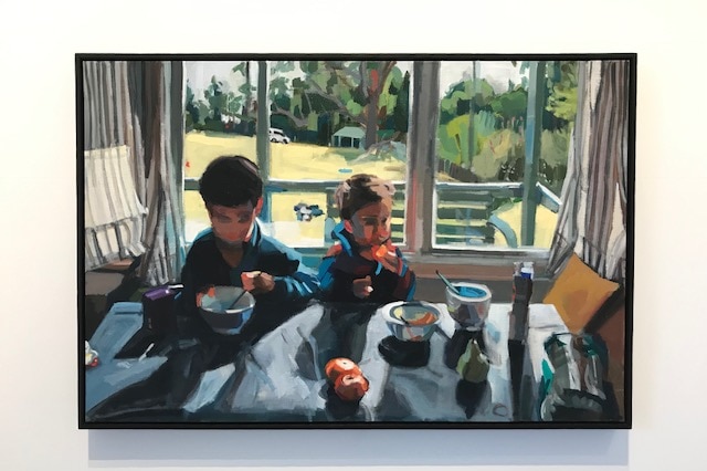 The painting hung on a gallery wall depicts two children eating breakfast in front of a large window.