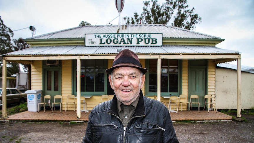 A man in a black leather jacket and pork pie hat outside a weatherboard pub.