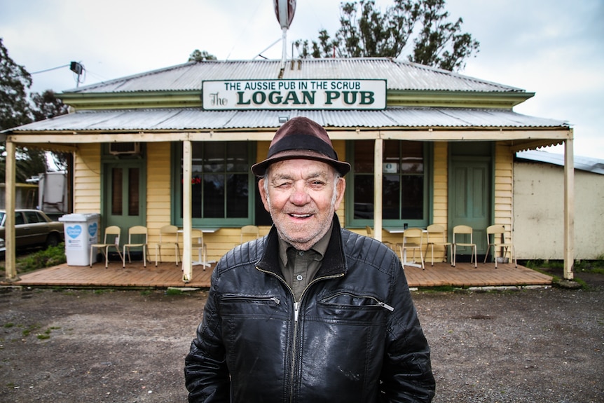 A man in a black leather jacket and pork pie hat outside a weatherboard pub.
