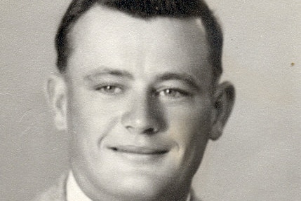 A black-and-white image of a young man with 1940's styled hair, a wide smile and in a jacket and tie posing for the camera.