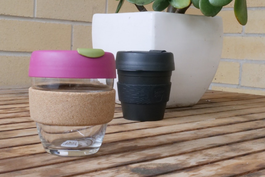 Two reusable cups sitting on a wooden table in front of a potplant, one is clear with a pink lid, other is all black