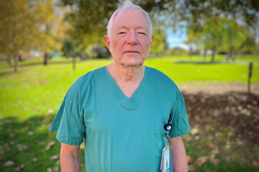 A man with light white hair wearing his green scrubs looks sternly at the camera in a park