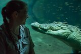 A silhouetted woman sits face to face with a crocodile on the other side of thick aquarium glass.