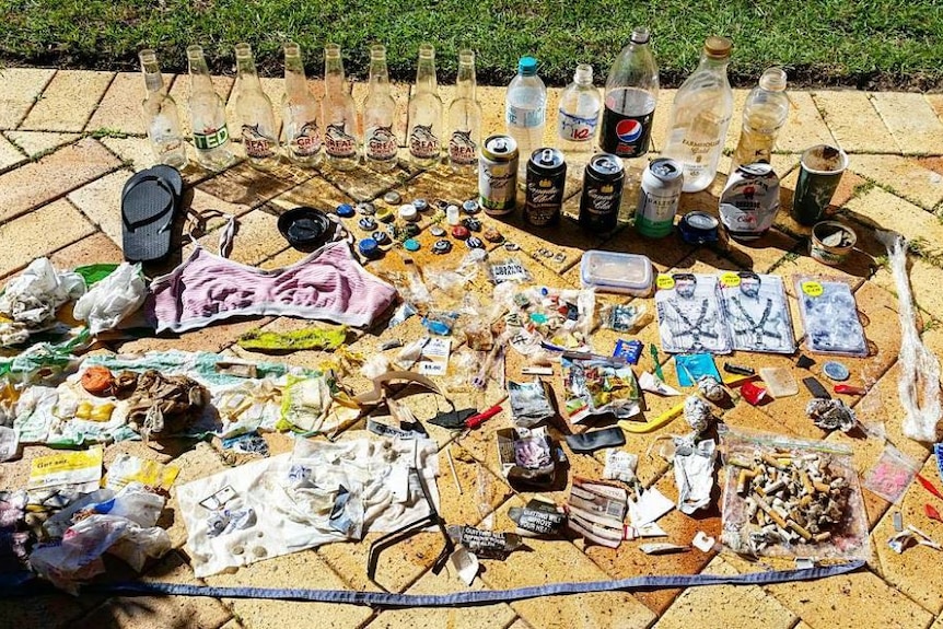 Empty drink containers, cigarette butts, a swimming top, a thong and other pieces of discarded rubbish lined up on pavers