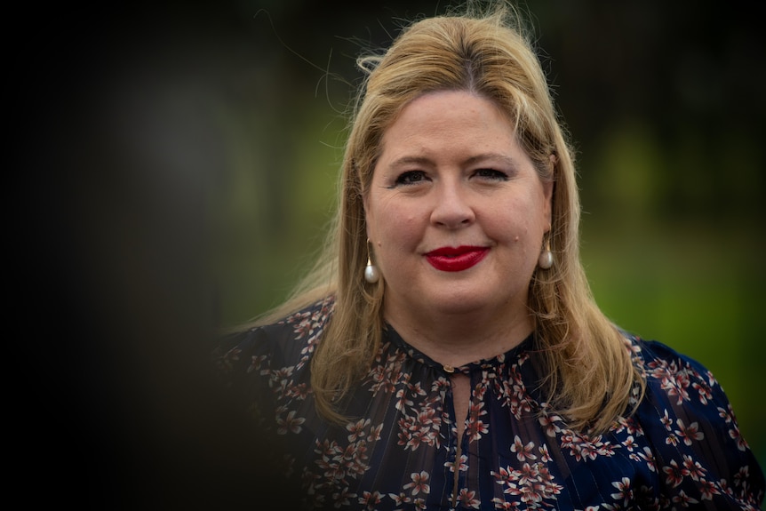 A woman with blonde hair smiles, red lipstick, black and pink printed blouse, pearl earings. 