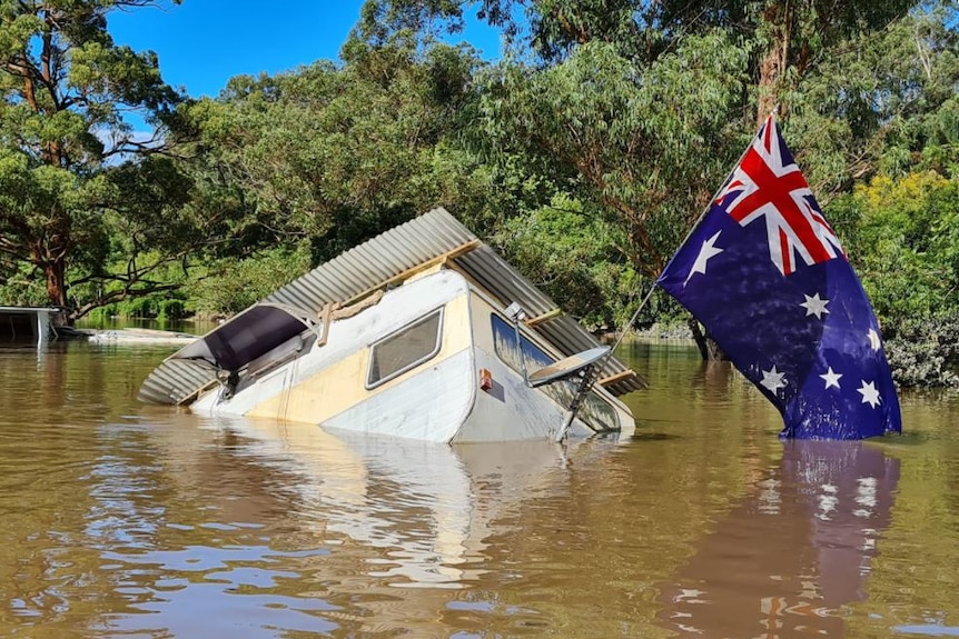 A caravan, with a huge Australian flag attached, is tipped over and partly submerged in brown floodwaters.