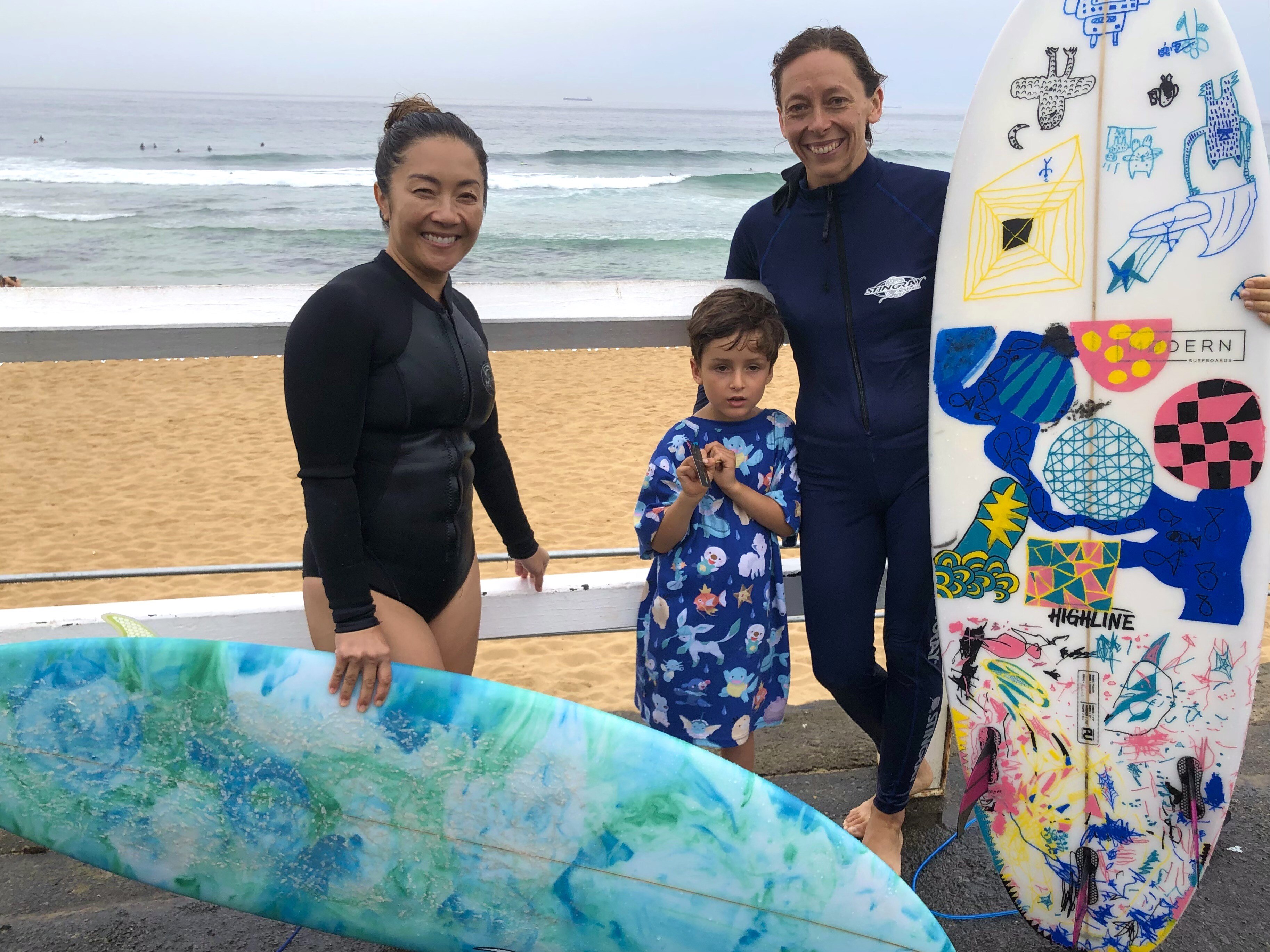 Two women stand in front of the surf with their surfboards. A young boy stands in between them.