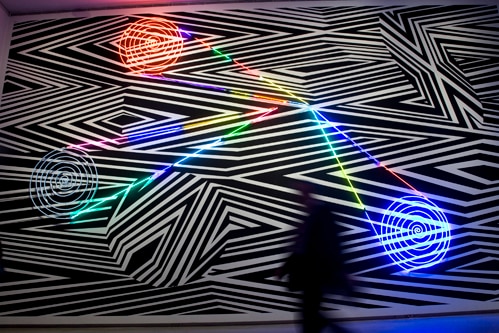 A geometric artwork on the wall of an art gallery which has black and white background with neon-coloured patterns.