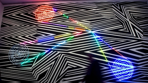 A geometric artwork on the wall of an art gallery which has black and white background with neon-coloured patterns.
