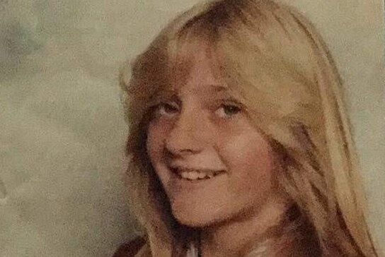 A smiling Jodie Larcombe wearing what appears to be a school uniform and a Farrah Fawcett hairstyle.