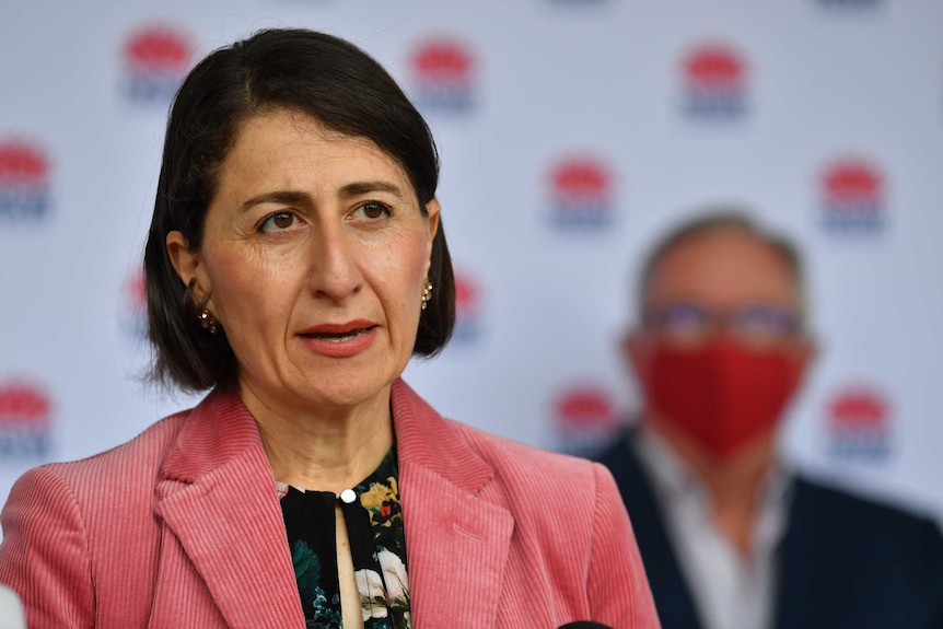 Premier Gladys Berejiklian wears a pink jacket and stand infront of Brad Hazzard, who is wearing a red face mask.
