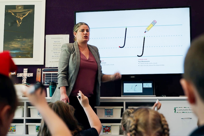 A teacher stands in front of a screen demonstrating how to draw the letter j.
