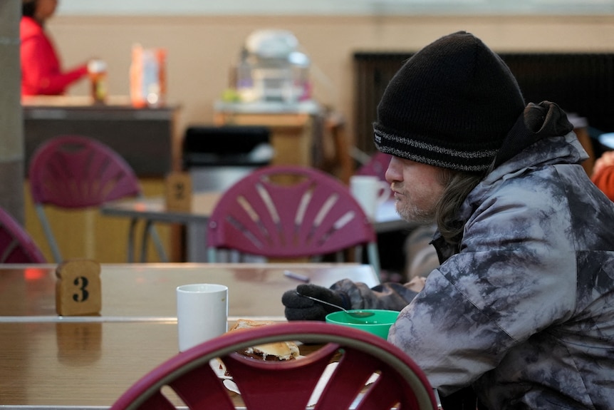A man wearing a beanie sits at a table eating breakfast.