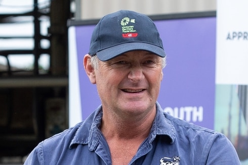 Older man with grey sideburns, caught mid-speech, wears a blue cap with some words, blue denim shirt, stands in front of signs.