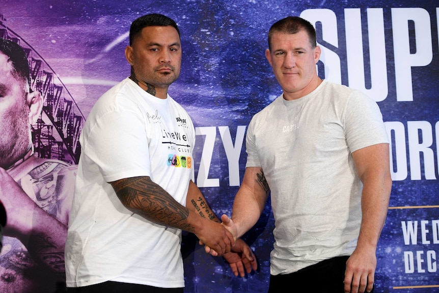 Mark Hunt and Paul Gallen stand and shake hands, both wearing white t-shirts in front of a blue background