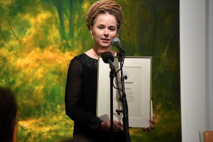 A woman speaks into a microphone while holding a framed award.