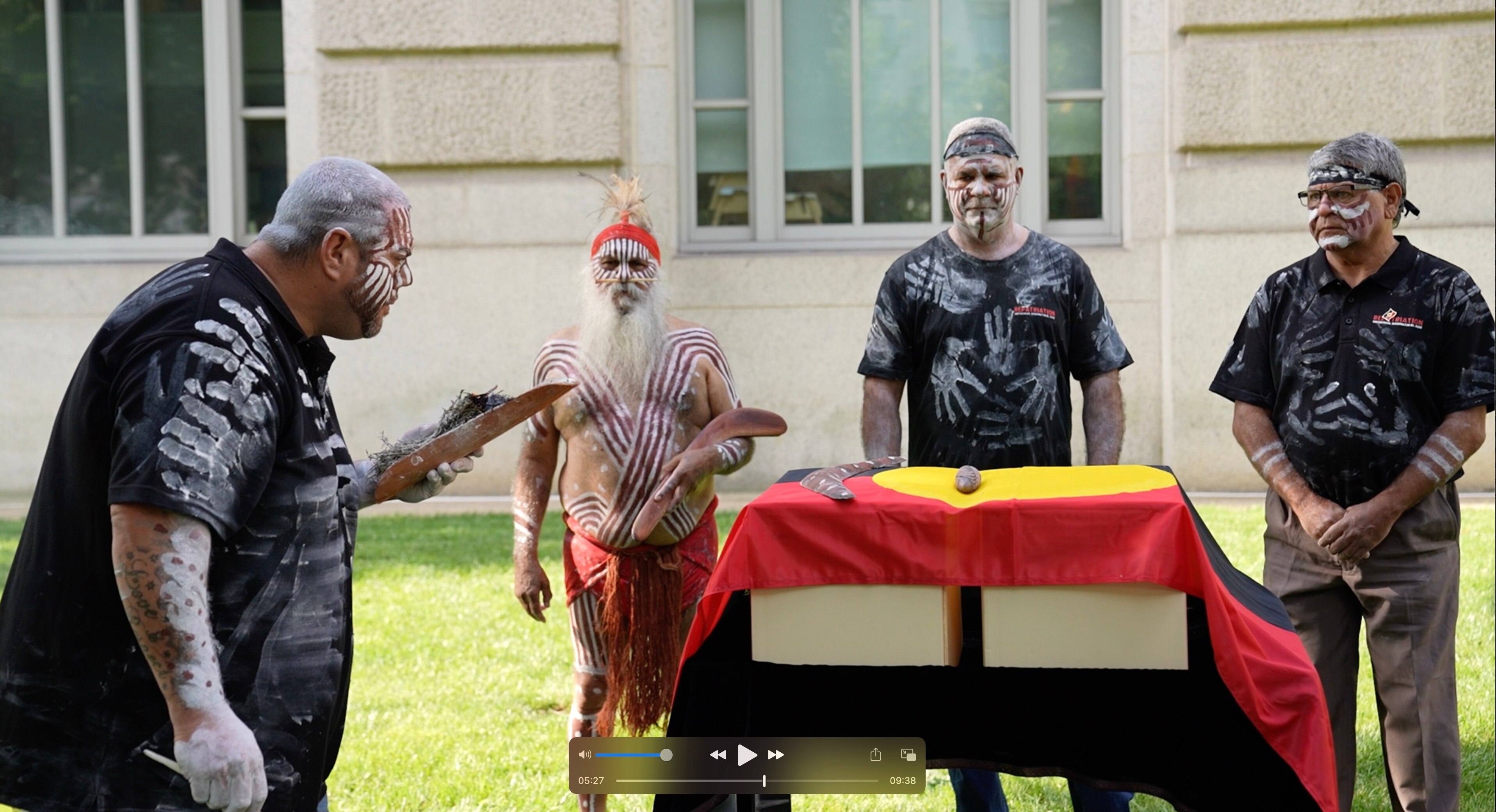 Four Indigenous men wearing traditional white and red paint stand around boxes draped in an Aboriginal flag
