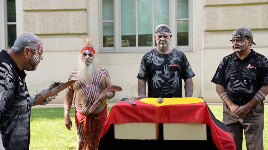 Four Indigenous men wearing traditional white and red paint stand around boxes draped in an Aboriginal flag