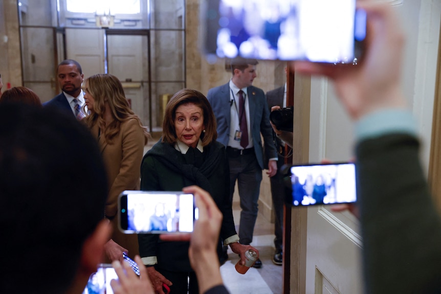 Nancy Pelosi walks past several reporters holding up their phones to record video. 