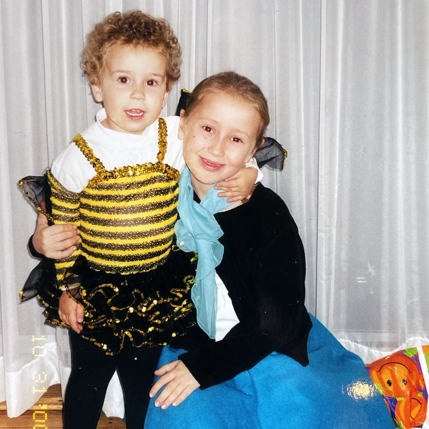 A toddler dressed in a bee costumer wraps her arm around an older girl's neck 