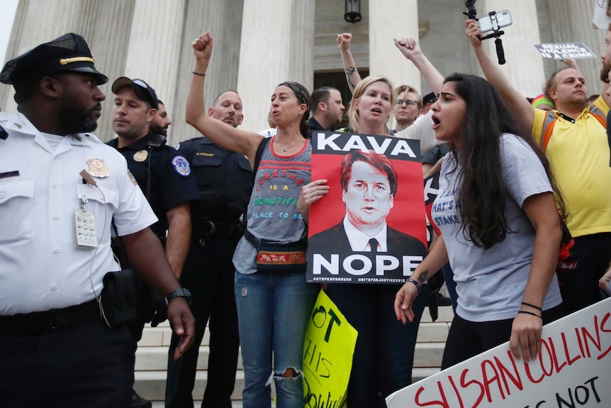 A police officer looks at a group of women standing outside the Capitol building, one holds a "KAVANOPE" sign