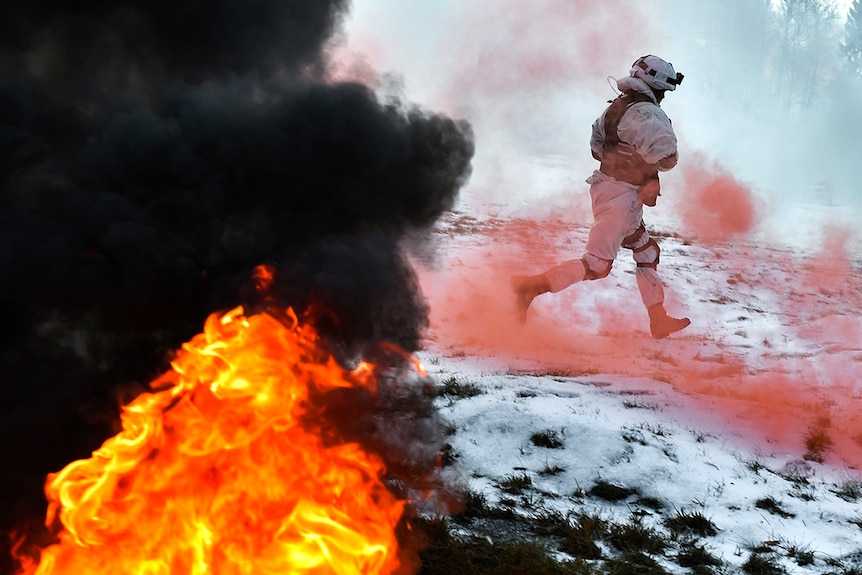 A Russian soldier skips past red smoke from a flare as a fire rages in the foreground