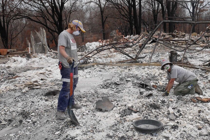 Two people, wearing face masks and grey shirts, sift through rubble after a bushfire