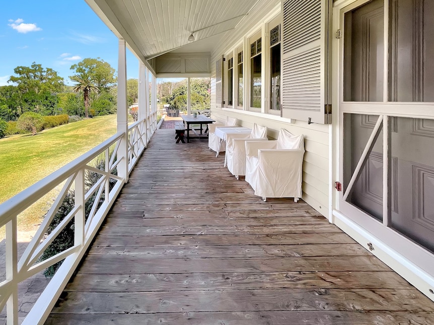 A wide wooden verandah with a rail along the side of a white house with windows. 