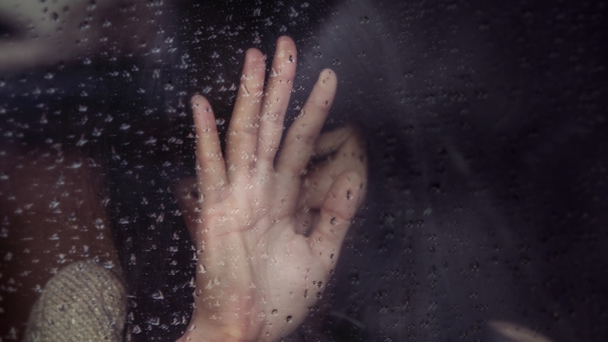 A Distressed woman with her hand on a pane of glass with rain droplets.